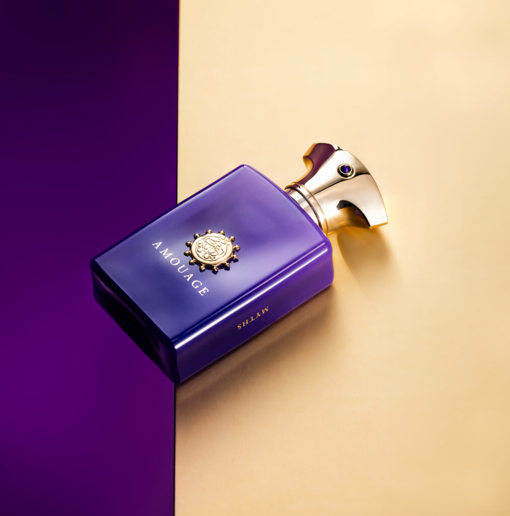Purple Amouage perfume bottle positioned diagonally across a geometric purple and golden background.