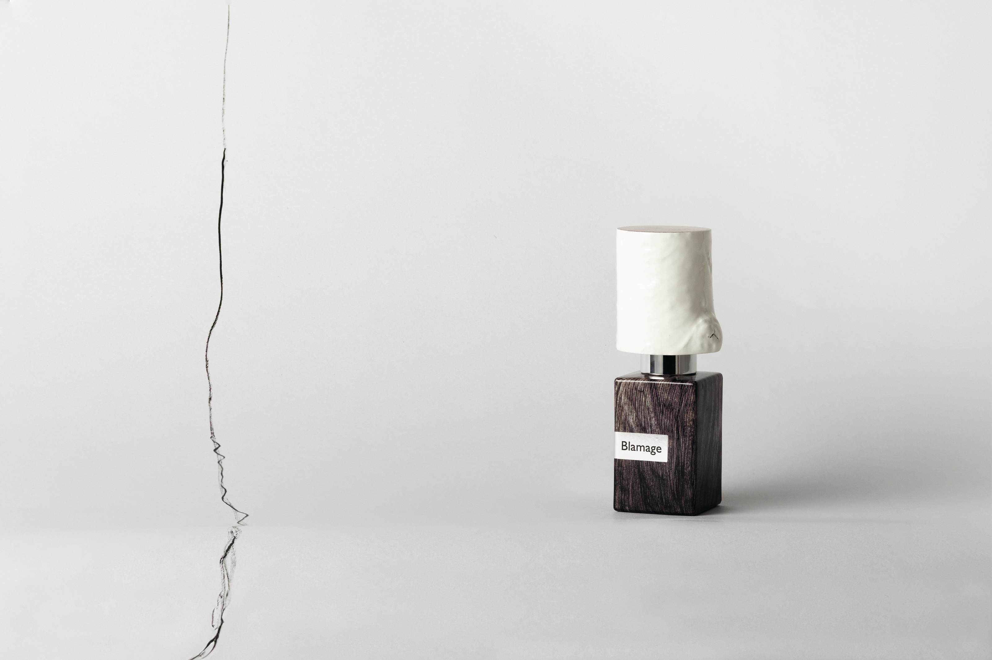 Nasomatto Blamage perfume bottle standing on a pale background, with a thin irregular dark line running vertically to its left.