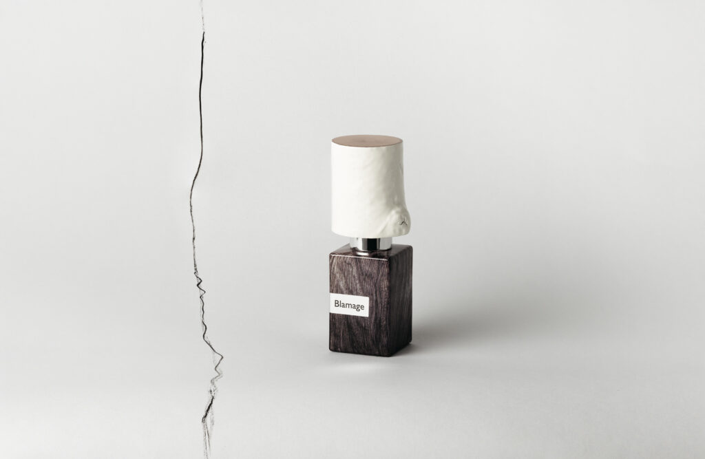 Nasomatto Blamage perfume bottle standing on a pale background, close to a thin irregular dark line running vertically to its left.