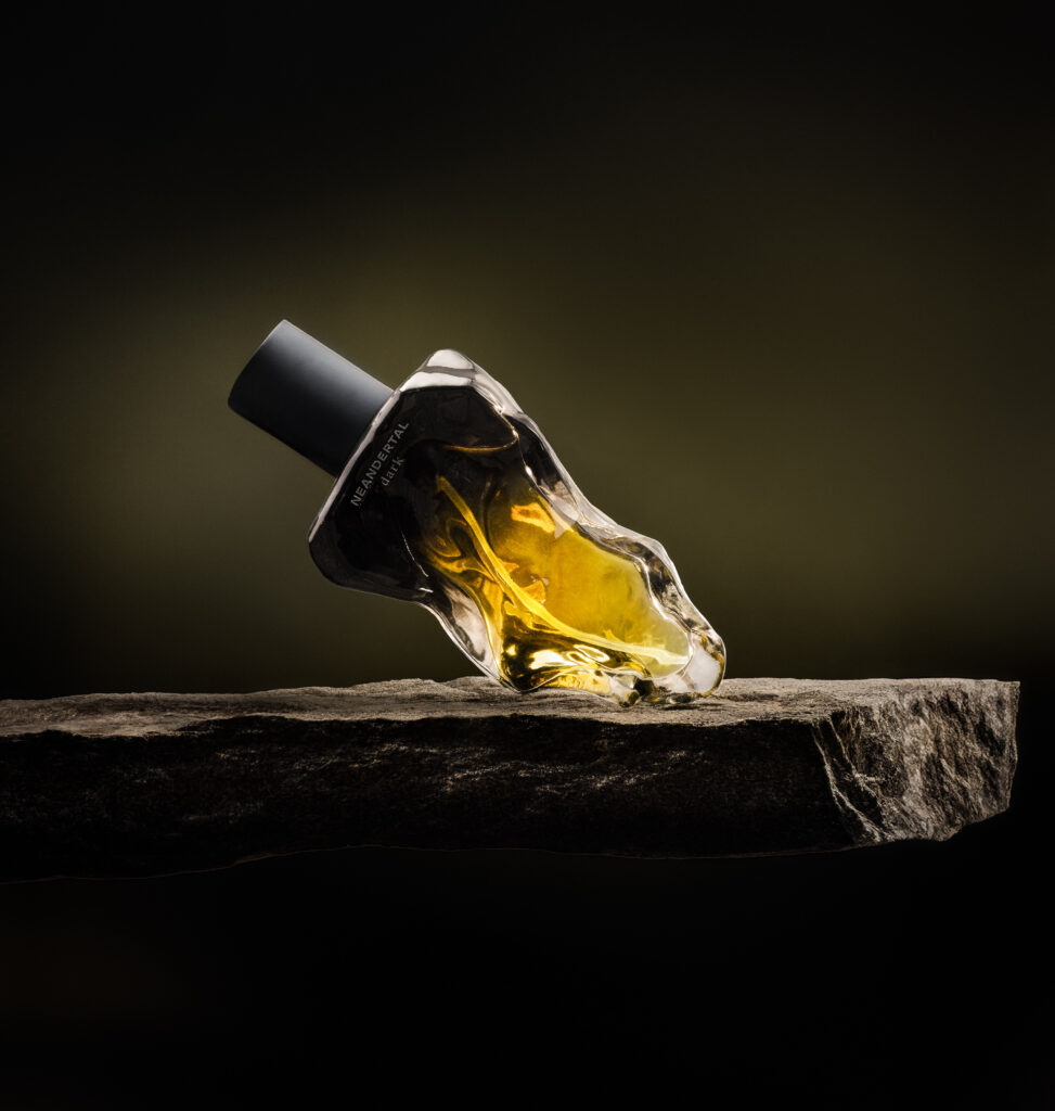 Neandertal Dark perfume bottle tilted on a rough stone slab, with illuminated amber glass against a dark olive background.