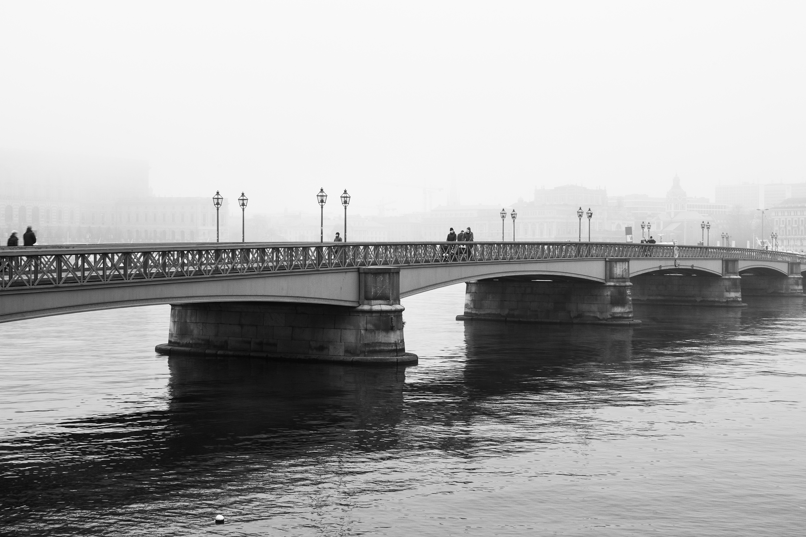 Black and white photograph of a bridge crossing the water on a foggy day, with pedestrians and distant buildings fading into the mist.