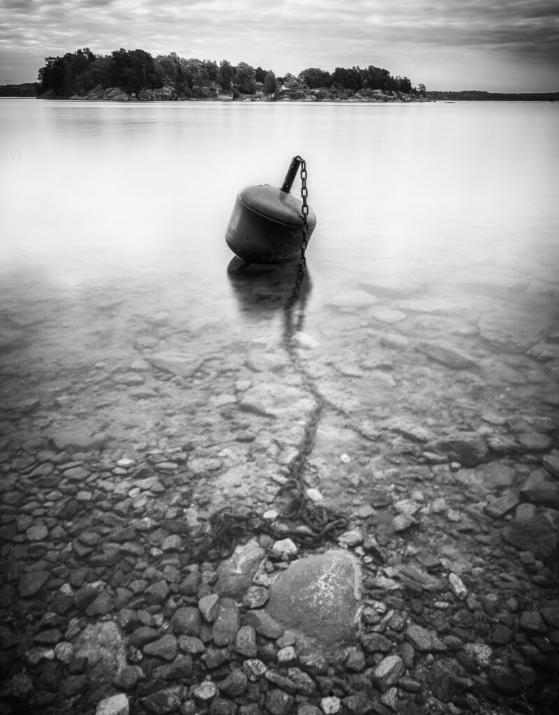 Black and white long-exposure photograph of a mooring buoy and its chain in shallow water, with a wooded island in the distance.