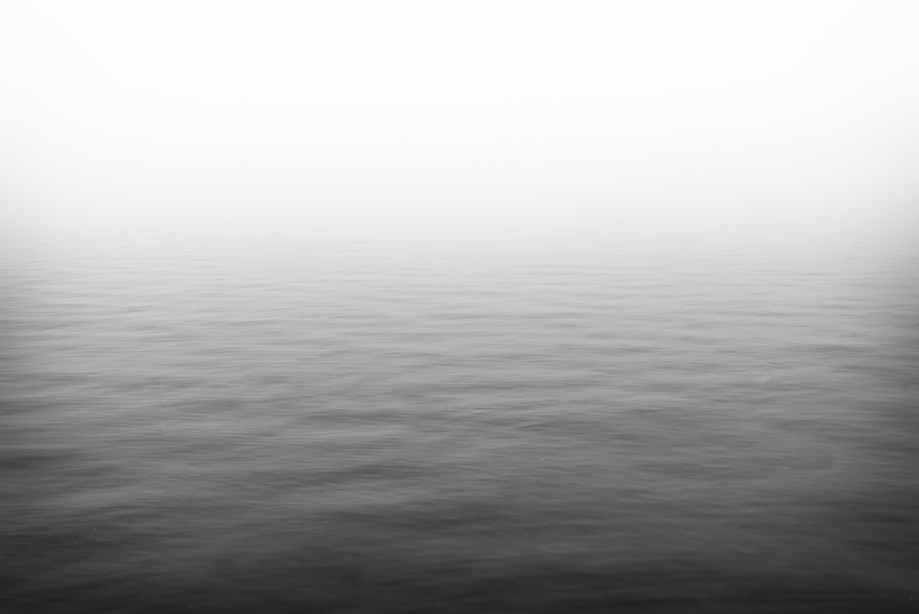 Minimalist black and white photograph of calm water fading into a distant horizon obscured by fog.