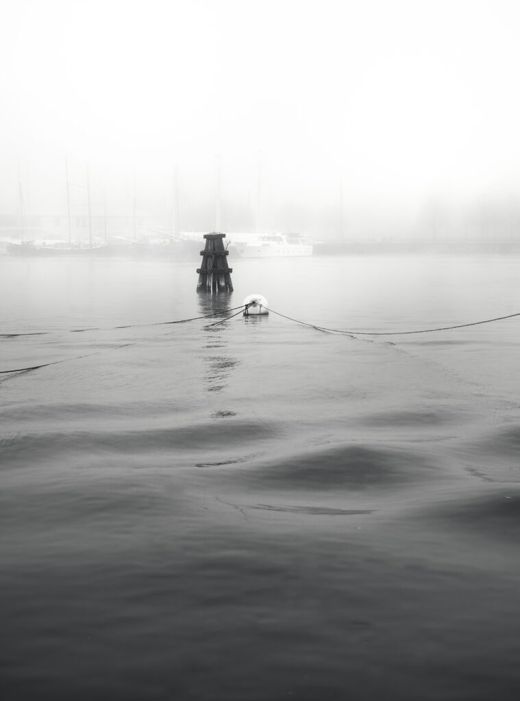 Mooring ropes stretching across calm water towards a small structure, with boats barely visible through the fog.