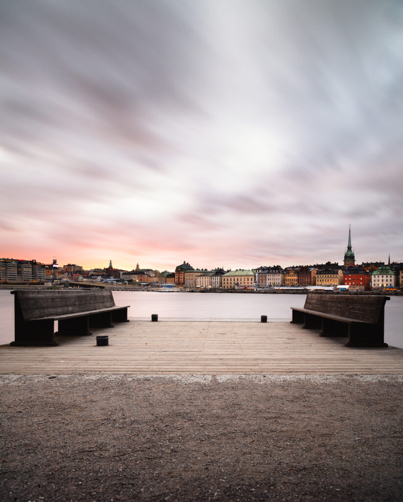 Two wooden benches facing Stockholm across calm water beneath a softly colored evening sky.