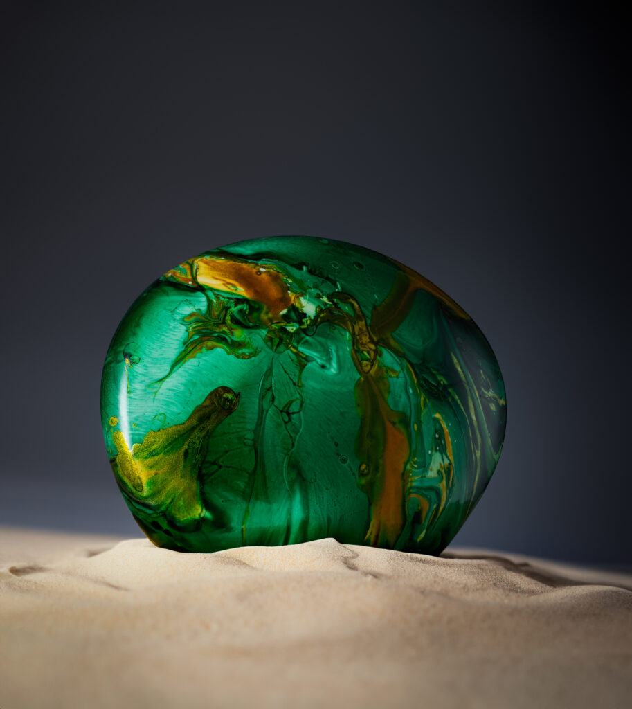 Rounded emerald-green perfume bottle nestled in pale sand against a dark background.