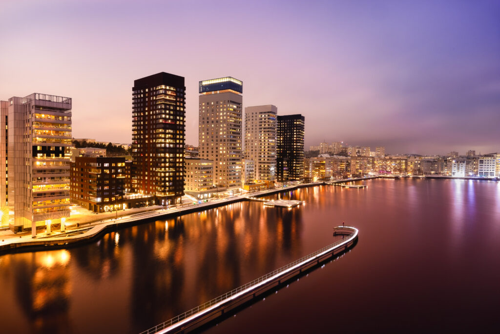 Modern waterfront buildings illuminated at dusk beneath a violet sky, with a curved pier extending into the water.