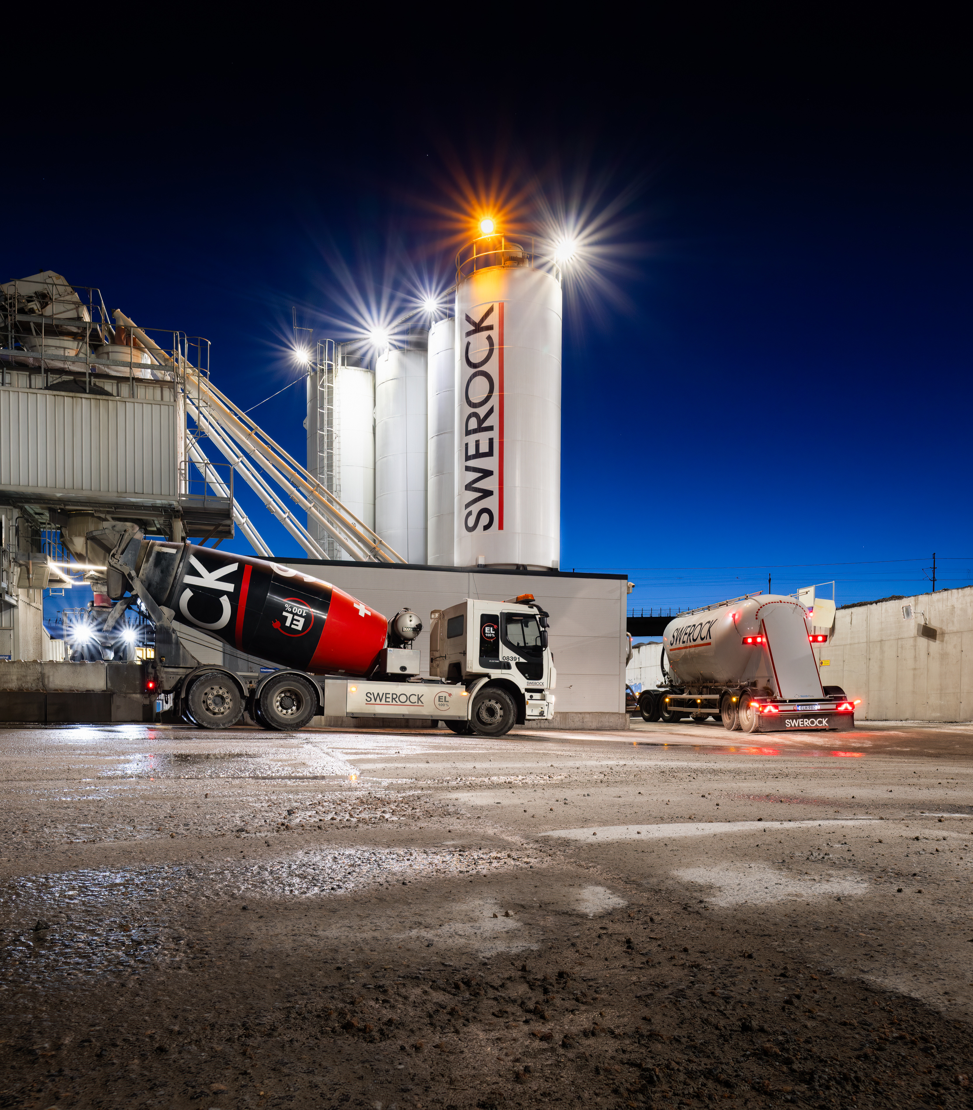 Two concrete mixer trucks at an illuminated concrete batching plant at night, photographed in dramatic low light.